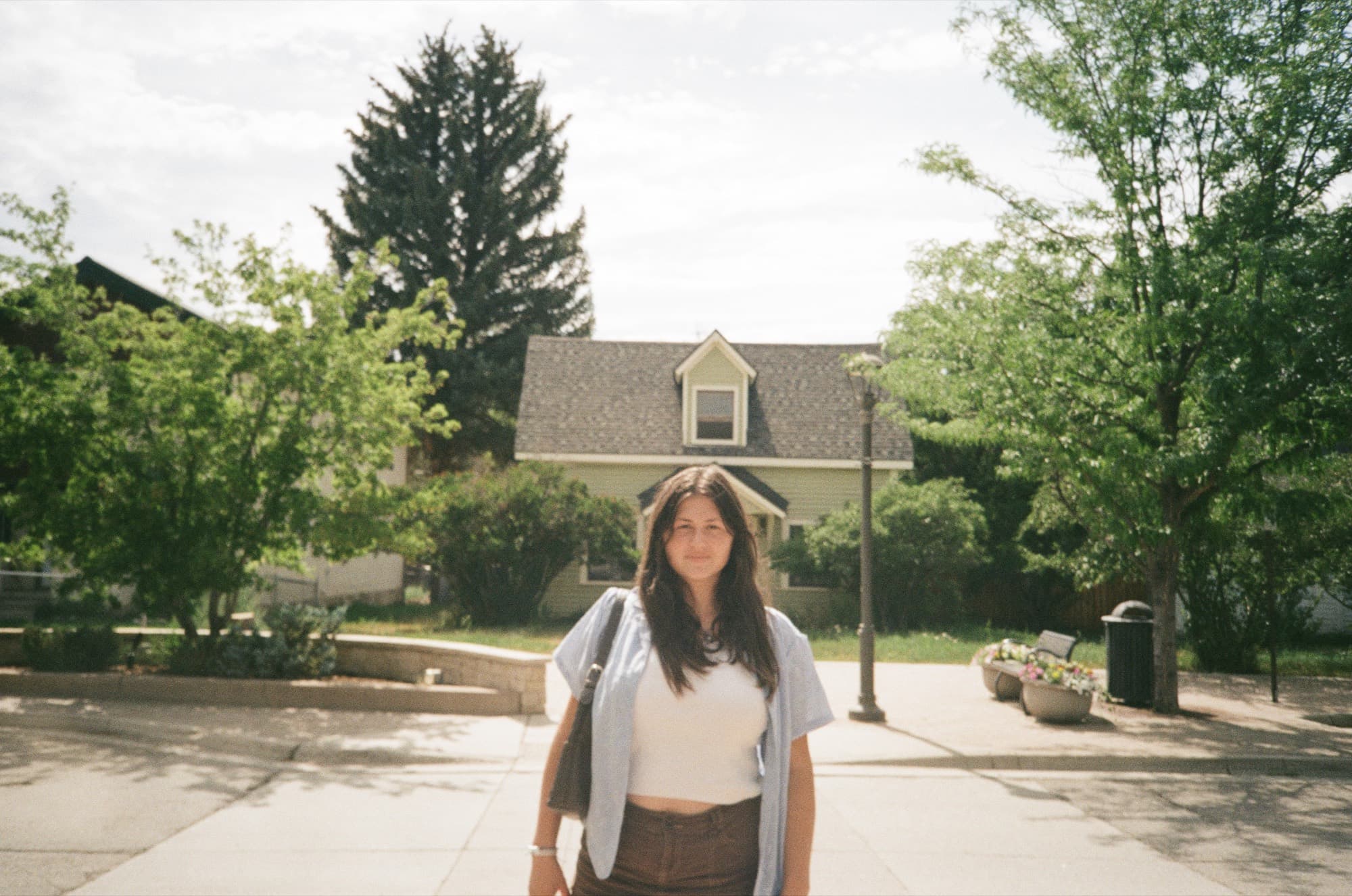 A woman standing on a sunlit pavement, a clapboard house and trees behind her
