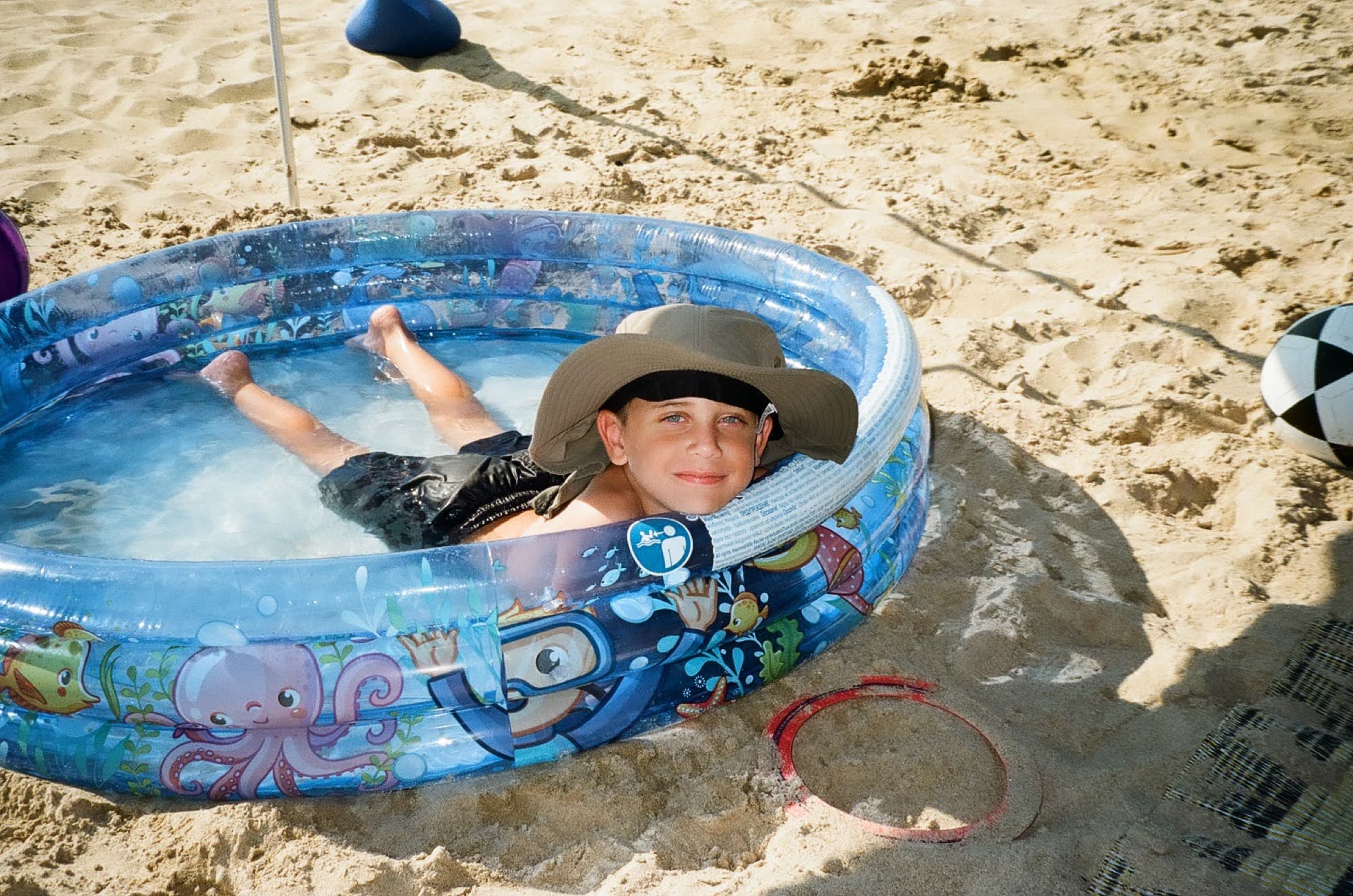A boy in a wide-brimmed hat lying back in an inflatable paddling pool on the sand