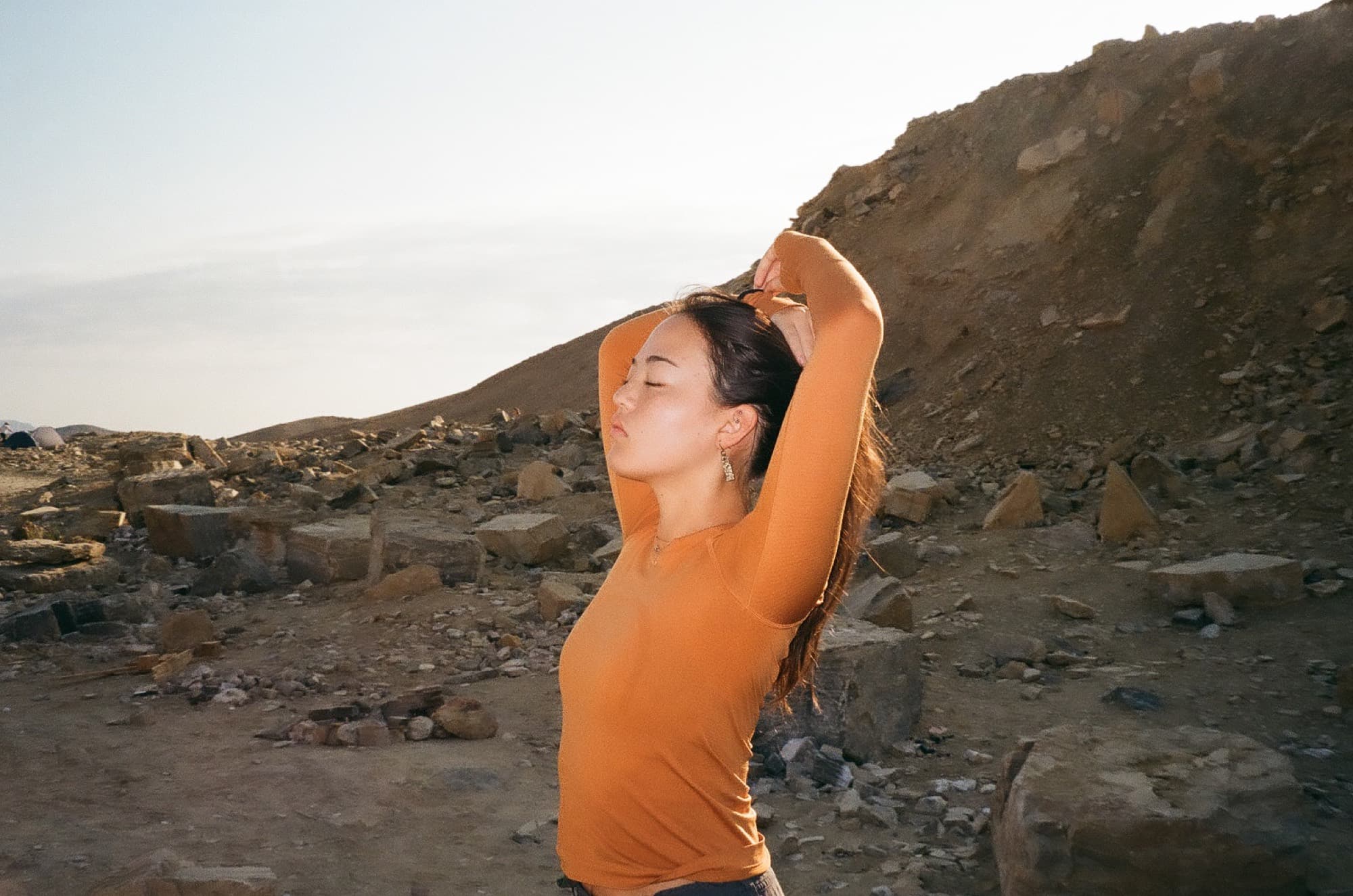 A woman in an orange top tying her hair back on a desert hillside at golden hour