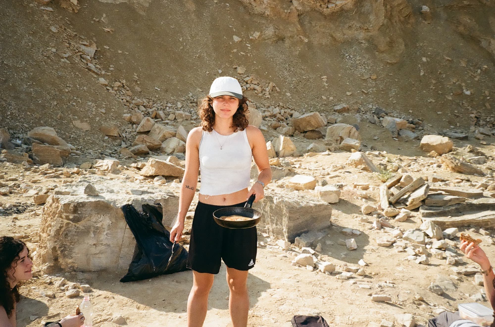A woman in a white cap holding a frying pan at a desert campsite