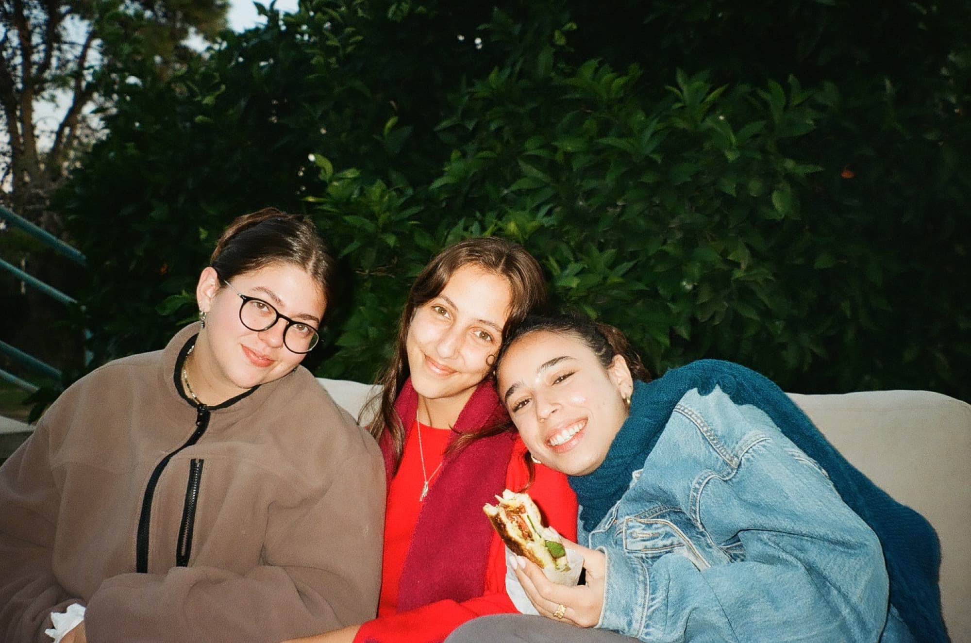 Three friends pressed together on a sofa outdoors in front of a dark hedge