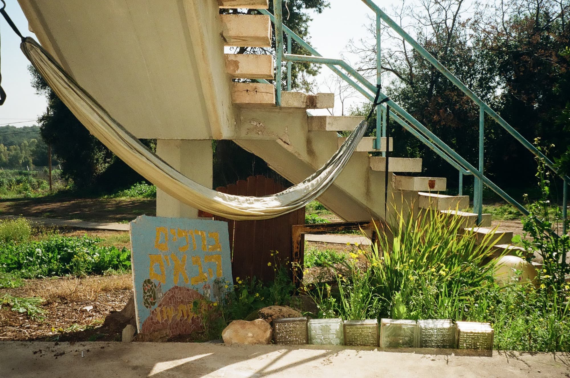 A hammock slung beneath a concrete staircase beside a hand-painted Hebrew sign