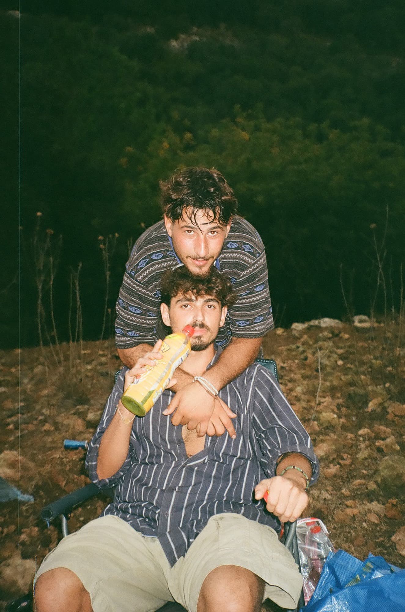 Two friends sharing a camping chair on a hillside at dusk