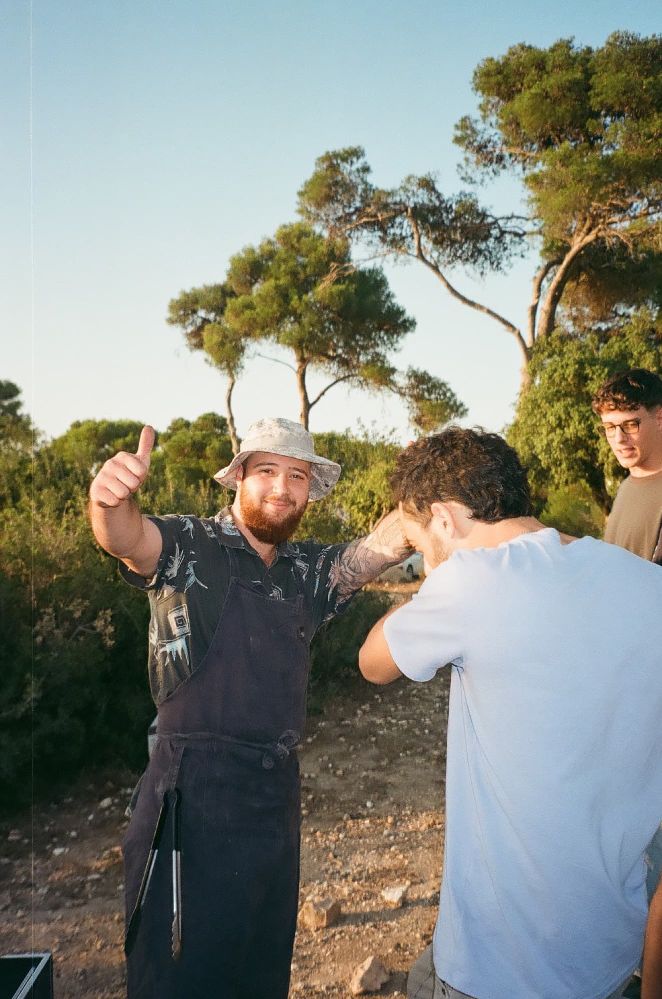 A cook in an apron giving a thumbs-up at an outdoor grill, pines behind