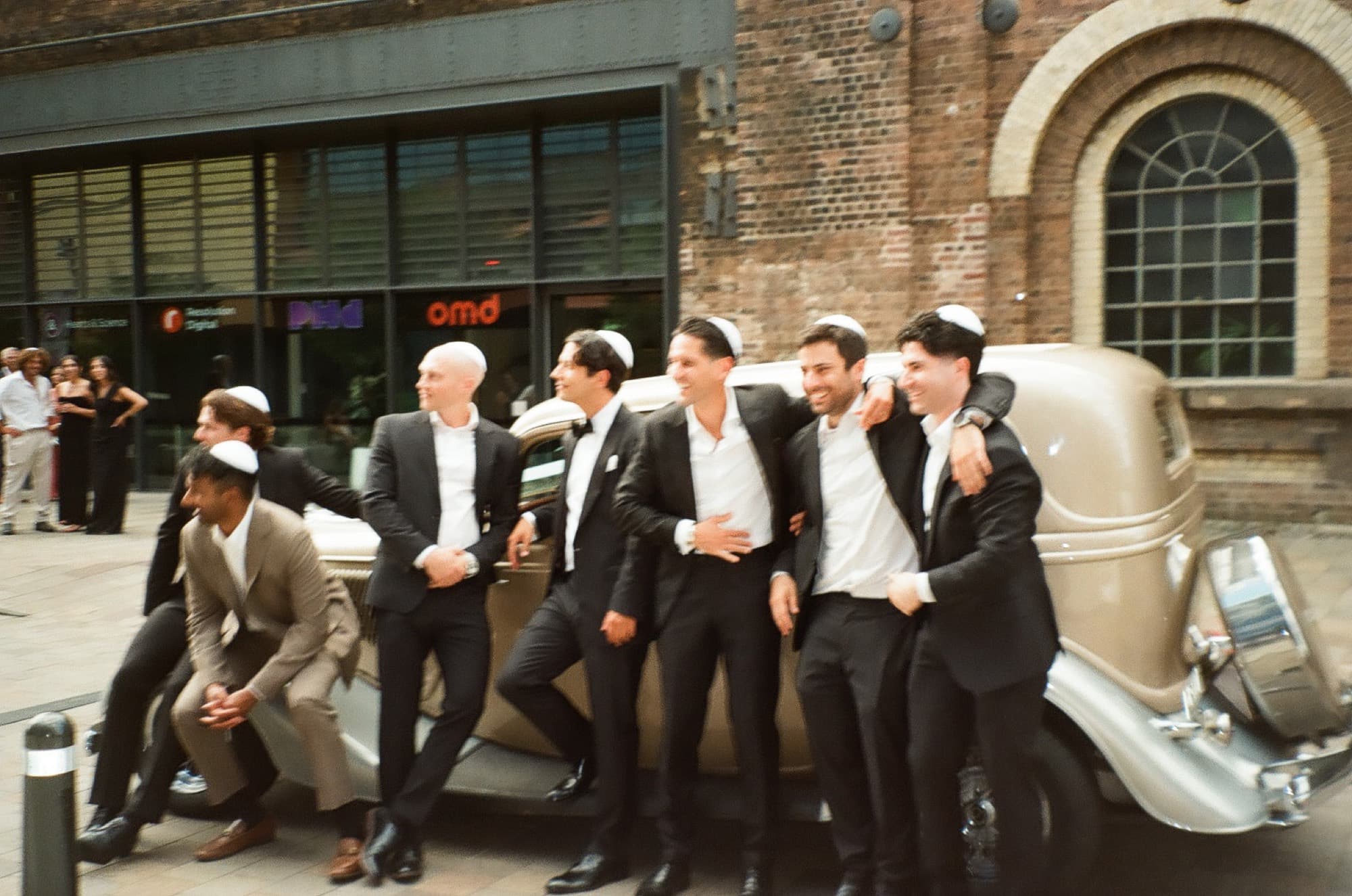 Groomsmen in kippot leaning against a vintage car outside a brick building