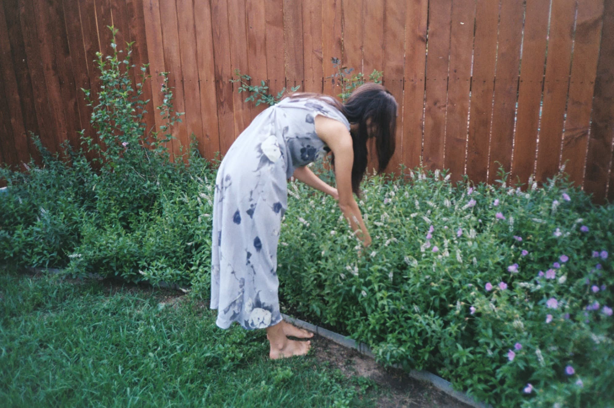 A woman in a long printed dress bending to touch flowers along a wooden fence