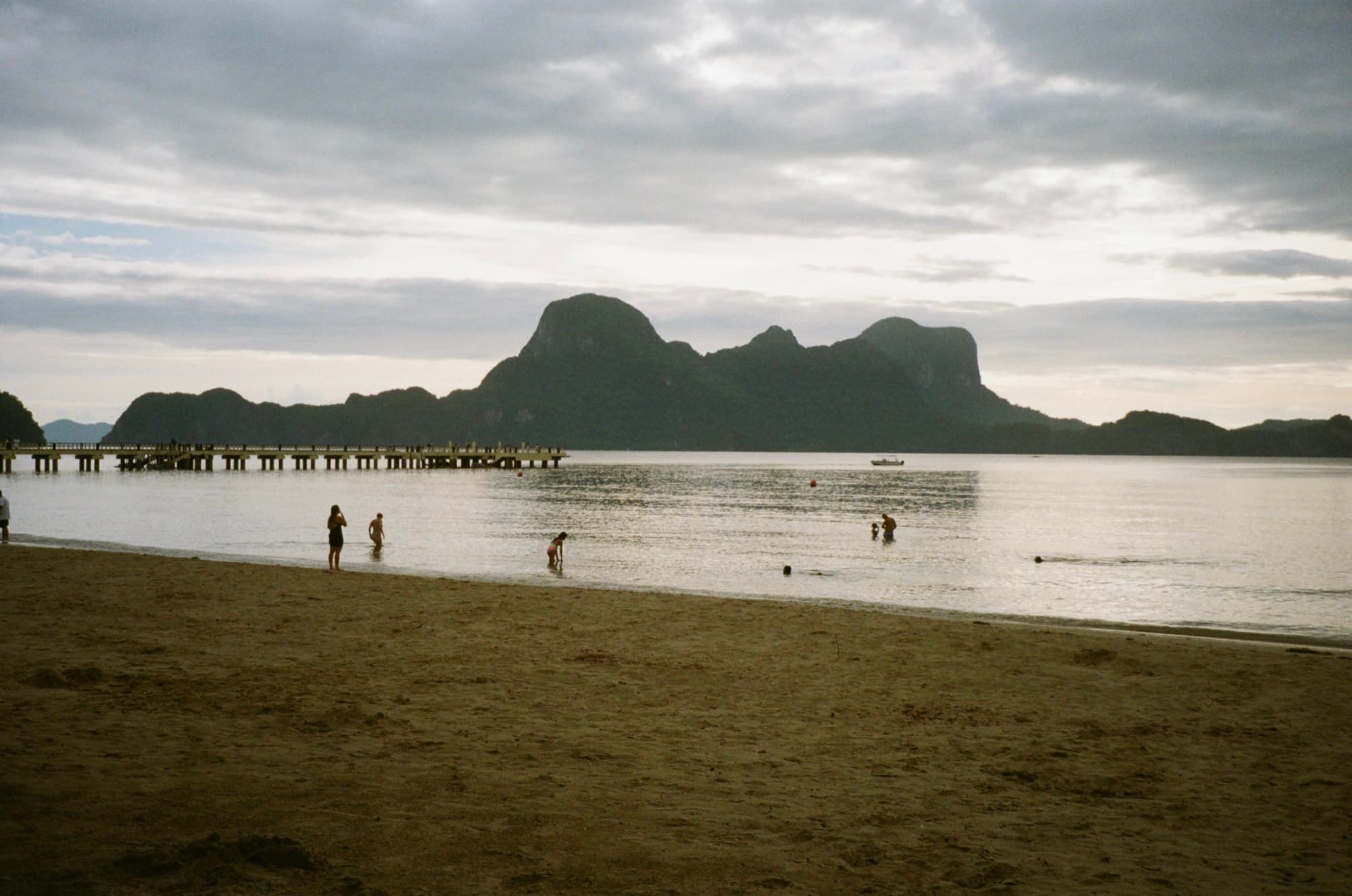 Swimmers in shallow water at dusk, karst headlands and a long pier across the bay